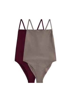 Donna in piedi fino alla vita in una piscina che indossa un costume intero grigio, con un muro di pietra e vegetazione sullo sfondo.