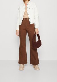 Chaqueta de mezclilla blanca, blusa de rayas, pantalones acampanados marrones y un pequeño bolso de mano marrón y esponjoso. Unas plataformas beige completan el conjunto.