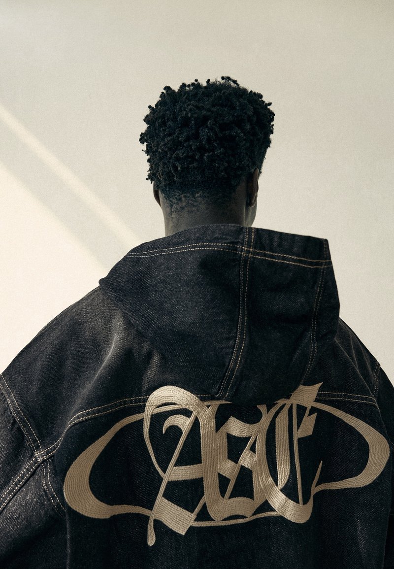 Personne aux cheveux courts et bouclés portant une veste en jean foncée avec un grand motif stylisé clair dans le dos, dos à la caméra.