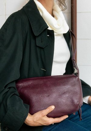 Borsa in pelle bordeaux con superficie texture, forma allungata e tracolla regolabile, tenuta vicino al corpo. Dettagli hardware lineari visibili.