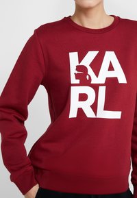 Bordeaux sweatshirt tillverkad av bomullsblandat material med stor vit text "KARL" och en siluettgrafik på framsidan. Långa ärmar, rundad halsringning.