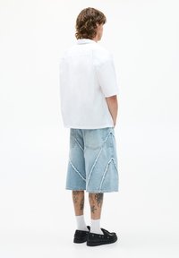 Camicia bianca a maniche corte con colletto, abbinata a pantaloni corti in denim blu chiaro con orli sfrangiati, caratterizzati da un motivo di cucitura geometrico e tasche. Scarpe nere.
