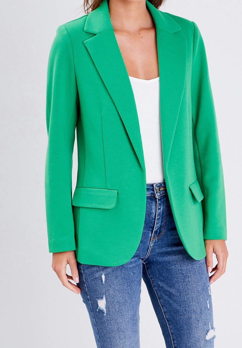 Femme portant un blazer vert vif sur un haut blanc et un jean bleu déchiré, debout devant un fond clair uni.
