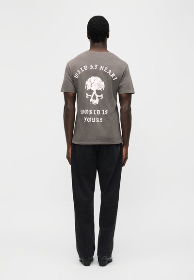 Άνδρας που φοράει γκρι μπλουζάκι με λευκό ξεφτισμένο κρανίο και κείμενο «WILD AT HEART» και «WORLD IS YOURS» στην πλάτη, συνδυασμένο με μαύρο παντελόνι.