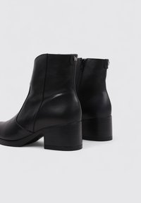 Botas de cuero negras hasta el tobillo con punta redondeada, tacón bajo y apilado, y cremallera lateral. Presenta un acento de logo en la parte trasera. Acabado suave.