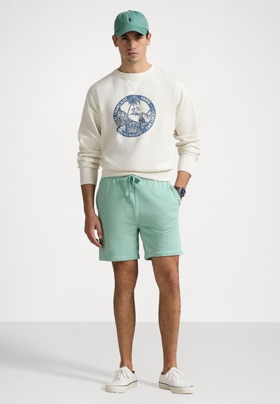 Lys kremgenser med blå grafikk, mintgrønne shorts og hvite sneakers. En matchende mintcaps fullfører antrekket.