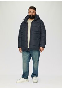 Marineblaue Steppjacke mit abnehmbarem Kragen, Reißverschluss vorne und dekorativen Taschen. Kombiniert mit hellblauen Jeans und weißen Sneakern.