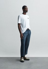 Moški stoji oblečen v belo srajco s kratkimi rokavi, modre skrajšane hlače, temne nogavice in dvobarvne mokasinke pred enostavno svetlo ozadje.