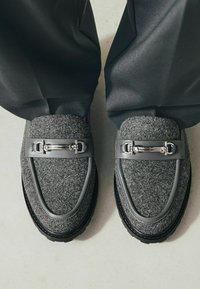 Grijze leren loafers met een soepel grijs leren bandje en zilveren gespdetails. Gevoerde bovenkant, zwarte rubberen zool, vanuit bovenaf bekeken.