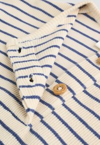 Gestreiftes Strickkleidungsstück in Creme mit marineblauen Linien. Verfügt über Holzknöpfe und einen strukturierten Stoff. Mit Kragen und Knopfleiste.