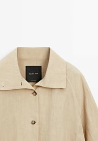 Beżowa lniana koszula z szpiczastym kołnierzykiem, z metką wewnątrz i dwoma guzikami z przodu. Gładka faktura i prosty design.