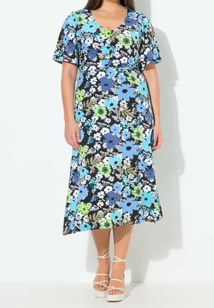 Femme portant une robe midi noire à imprimé floral bleu, vert et blanc avec des manches courtes, associée à des sandales à plateforme blanches à brides.