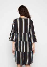 Černé šaty s vertikálními pruhy v modré, žluté a hnědé barvě. Mají vrstvený design a rozšířené rukávy. Zobrazeno ze zadní strany.