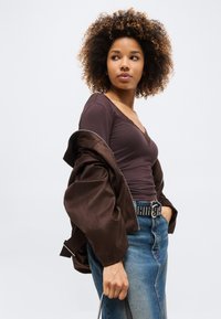 Νεαρή γυναίκα με σγουρά μαλλιά, φοράει σκούρο μωβ τοπ, καφέ μπουφάν εκτός ώμων, φούστα από τζιν και μαύρη ζώνη με καρφιά, κοιτάζει πλάγια.