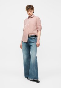 Άτομο που φοράει ανοιχτό ροζ πουκάμισο με κουμπιά και μανίκια γυρισμένα, φαρδιά ξεβαμμένα μπλε τζιν, μαύρα παπούτσια και μαύρη ζώνη.