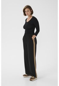 Sieviete, kas valkā melnu garām piedurknēm topu, melnas platajiem bikšu kājiņām ar brūnā raksta svītru sānos un brūnas kedas, stāv ar rokām kabatās.