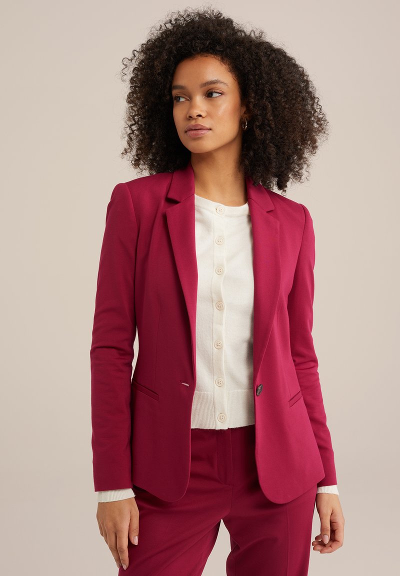 Fuchsia blazer med en knapp, sned lapel och två sidofickor, lagd över en vit knäpp cardigan, bärs med matchande byxor.