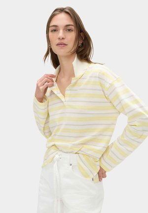 Vrouw draagt een geel-wit gestreept poloshirt met lange mouwen en witte hoge taille broek, hand past de kraag aan, neutrale achtergrond.