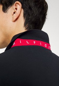 Läheltä kuvattu henkilön kaula ja korva, jolla on päällään musta kauluspaita, jossa on punainen nauha, joka näyttää osittain valkoisia kirjaimia kauluksessa.