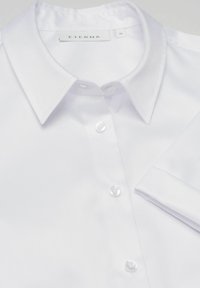 Chemise blanche en tissu lisse, dotée d'un col classique, d'une patte de boutonnage avec quatre boutons et d'un poignet replié. Étiquette de marque visible.