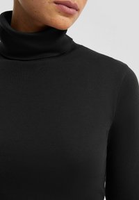 Crna turtleneck majica izrađena od glatkog, rastezljivog materijala. Ima visok ovratnik i dugačke rukave. Prikaz u krupnom planu ističe teksturu i kroj.