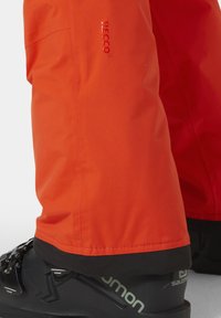 Jasně oranžové voděodolné kalhoty s černým lemem, logem RECCO, jsou doplněny černými lyžařskými botami s logem Salomon.