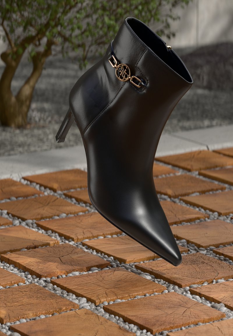 Černé kožené kotníkové boty s špičatou špičkou a malým podpatkem. Vyznačují se zlatým řetízkovým detailem na kotníku. Umístěny na texturovaném kamenném povrchu.