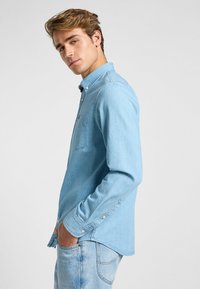 Ljusblå denimskjorta med långa ärmar, knapplistkrage, en bröstficka och manschetter med knappar. Bärs med ljusblå denimbyxor.