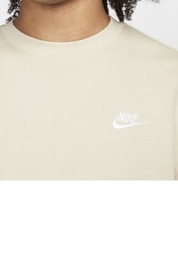 Beige bomullströja med rund halsringning som har en liten vit Nike-logga på vänster bröstområde, slät textur och ribbad halsringning.