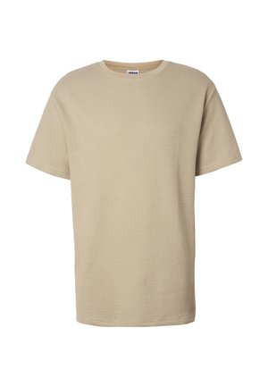 Jeune homme aux cheveux bouclés portant un t-shirt beige à manches courtes texturé et un jean foncé, debout devant un fond clair uni.