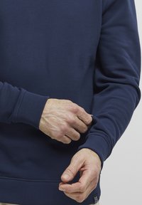 Sweatshirt blu navy con polsini e orlo a costine, caratterizzato da una struttura tessile e un design aderente. Il modello regola la manica vicino al polso con le mani.