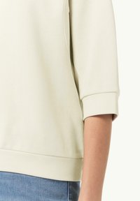 Close-up van een persoon die een crème-kleurige top met lange mouwen en omgeslagen manchetten draagt, gecombineerd met blauwe spijkerbroek.