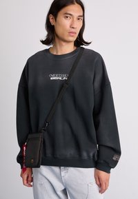 Felpa nera con testo bianco, vestibilità oversize, maniche a costine, abbinata a una piccola borsa nera a tracolla, indossata sopra dei jeans chiari.