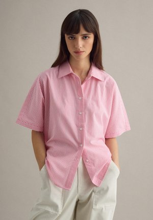 Young woman with straight brown hair wearing a pink and white vertical striped short-sleeve button-up shirt and light beige pants with hands in pockets.