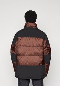 Giubbotto imbottito con un design a blocchi di colore in pannelli bronzeo, marrone e nero. Realizzato in materiale lucido e trapuntato con un colletto alto.