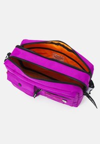 Fialová látková taška se dvěma zipovými přihrádkami, oranžovým vnitřkem, černými zipovými akcenty a štítkem. Malá přední kapsa se zipem.