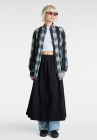 Camisa de cuadros en tonos de verde y negro, top corto, falda larga negra y pantalones de pierna ancha en azul claro; accessorios con gafas de sol.