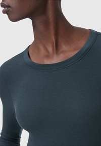 Top a maniche lunghe a coste in misto cotone color teal scuro, con scollatura rotonda e una texture liscia, che evidenzia la elasticità e la vestibilità del tessuto.
