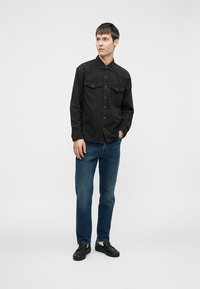 Svart skjorte med knepping og to brystlommer, kombinert med blå jeans og svarte sko. Modellen står mot en ensfarget lys bakgrunn.