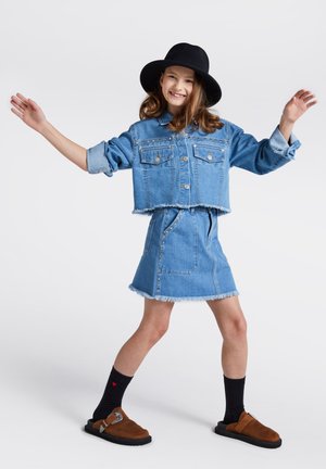 Dżinsowa kurtka o kroju crop z zapięciem na guziki i postrzępionym dołem, zestawiona z pasującą spódnicą o postrzępionych brzegach. Brązowe klapki i czarne skarpetki z wzorem w serca.