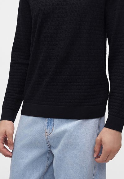 Personne portant un haut noir texturé à manches longues avec un motif de texte subtil et un jean denim bleu clair, coupé à la taille et aux cuisses.