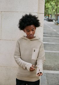Hoodie tricoté beige avec un design texturé, poche kangourou et trous pour les pouces. Les poignets côtelés et une poche zippée sur la poitrine ajoutent des détails.