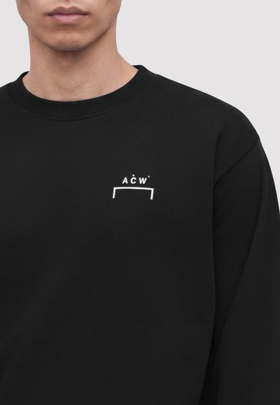 Κοντινό πλάνο ενός ατόμου που φοράει μαύρο φούτερ με λευκό λογότυπο "A C W" και οριζόντια γραμμή στην αριστερή περιοχή του στήθους.