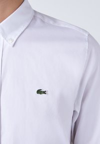 Chemise blanche à manches longues en coton, arborant un logo de crocodile brodé en vert sur la poitrine gauche. Texture lisse avec un col.