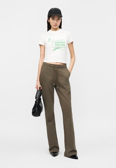 Γυναίκα μοντέλο που φοράει λευκό κοντό μπλουζάκι με πράσινο κείμενο "Döner House", ελιάς χρώματος παντελόνι με κορδόνι, μαύρο καπέλο, μαύρα μυτερά παπούτσια και κρατάει μαύρη τσάντα.