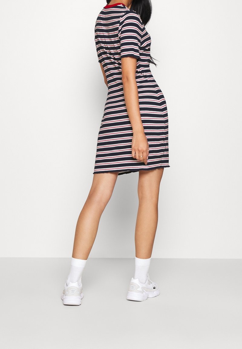 Robe rayée en bleu marine, rouge et blanc, avec des manches courtes et une silhouette ajustée. Portée avec des baskets blanches et des chaussettes courtes.