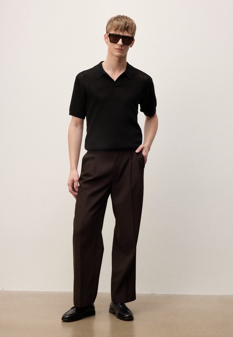 Jeune homme portant un polo en maille à manches courtes noir, un pantalon large marron foncé, des chaussures en cuir noires et de grandes lunettes de soleil noires, debout contre un mur uni.