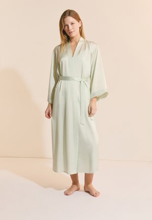 Kobieta stojąca boso, ubrana w długi, jasnozielony satynowy szlafrok z szerokimi rękawami i przewiązanym paskiem w talii.
