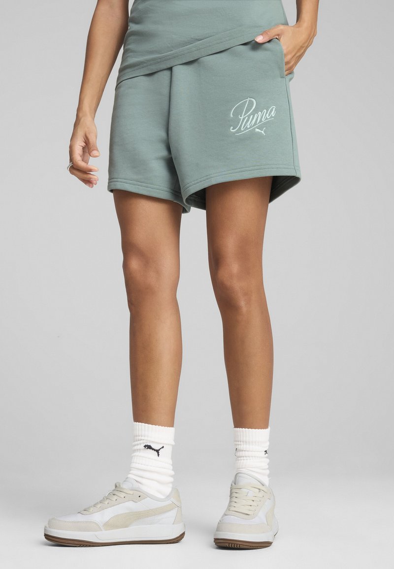 Pantalones cortos de sudadera suaves en verde menta con un corte holgado y un logo blanco de "Puma". Combinados con zapatillas ligeras y calcetas blancas de canalé.