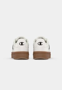 White sneakers with a low-top design, featuring a brown rubber sole, black logo accent, and textured heel detail.
